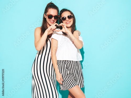 Two young beautiful smiling brunette hipster female in trendy summer clothes. Sexy carefree women posing near blue wall. Positive models having fun. Cheerful and happy. In sunglasses. Make heart sign