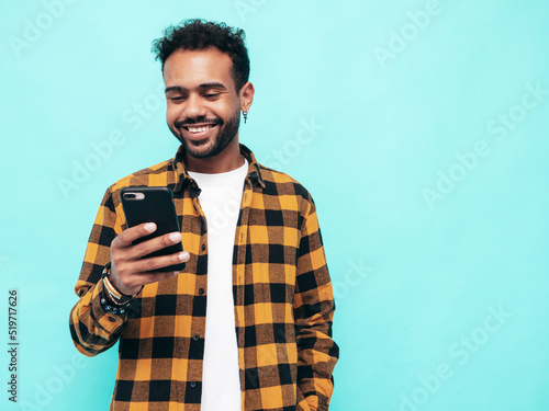 Handsome smiling hipster model. Sexy unshaven man dressed in yellow summer shirt and jeans clothes. Fashion male posing near blue wall. Holding smartphone. Looking at cellphone screen. Using apps