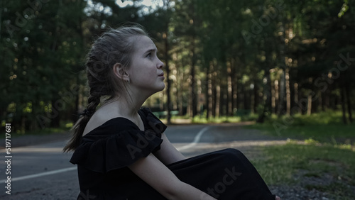 Lost schoolgirl sits on dark forest road. Teenager wearing black dress cries sitting alone on grass. Teen girl wants to find way to home among trees closeup