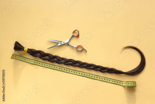 Long brown braid with scissors and tape measure on yellow background, donation of haircut, haircut selling service, material of natural hair for model hairstylish extension, top view