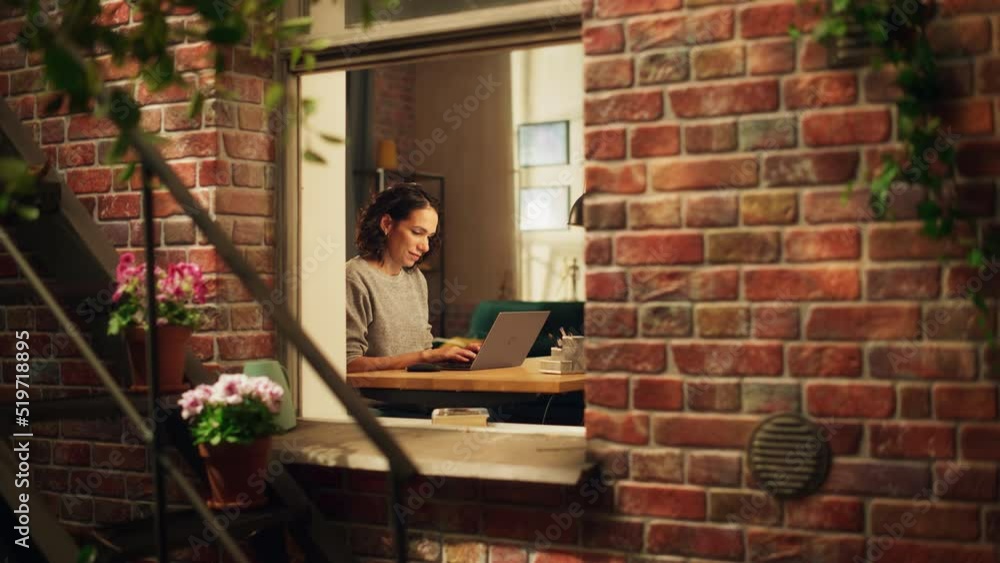 Hispanic Woman Using Laptop Computer, Sitting in Living Room im Her Apartment. Professional Female Working Remotely Online, Planning, Brainstorming New Project. Shot Into the Window From Outside. Hispanic Woman Using Laptop Computer, Sitting in Living Room im Her Apartment. Professional Female Working Remotely Online, Planning, Brainstorming New Project. Shot Into the Window From Outside.