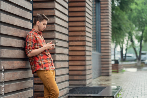 a young man in a plaid shirt stands leaning against the wall and looks at the phone