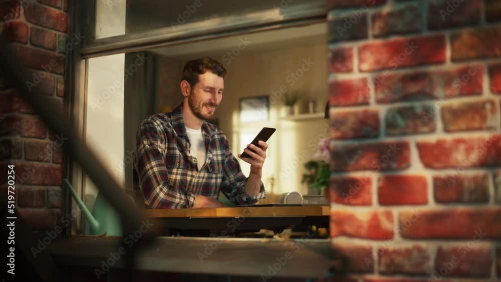 Happy Smiling Man Sitting Behind Desk at Home Office and Using Smartphone. Handsome Man Browsing Through Internet, Checking Videos on Social Media, Shopping Online. View From Outdoors into Window. Happy Smiling Man Sitting Behind Desk at Home Office and Using Smartphone. Handsome Man Browsing Through Internet, Checking Videos on Social Media, Shopping Online. View From Outdoors into Window.