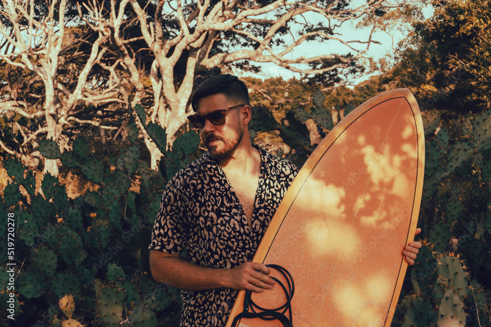 Surfer z deską surfingową, idący wybrzeżem na naturalnym tle, drzewa i kaktusy.