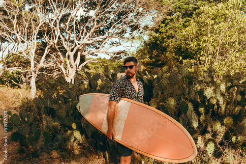 Obraz na płótnie Surfer z deską surfingową, idący wybrzeżem na naturalnym tle, drzewa i kaktusy. w salonie