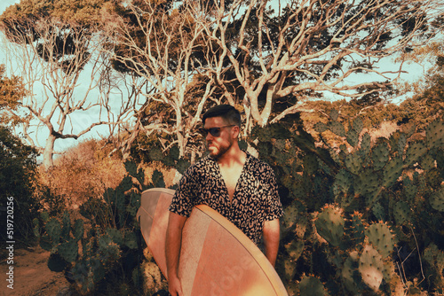 Obraz na płótnie Surfer z deską surfingową, idący wybrzeżem na naturalnym tle, drzewa i kaktusy. w salonie
