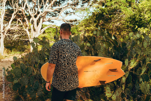Surfer z deską surfingową, idący wybrzeżem na naturalnym tle, drzewa i kaktusy.