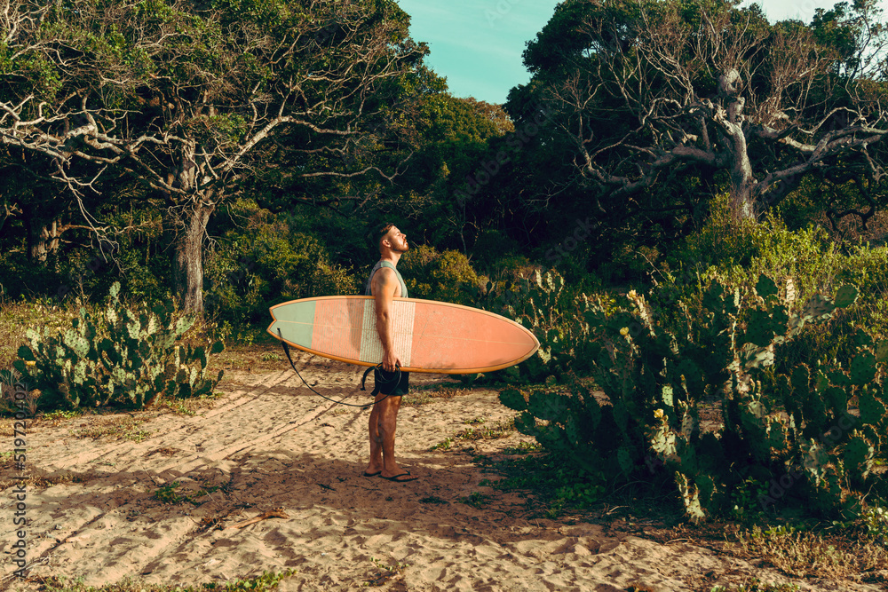 Mężczyzna surfer z deską surfingową, idący wybrzeżem na tle jungle, aktywny wypoczynek.