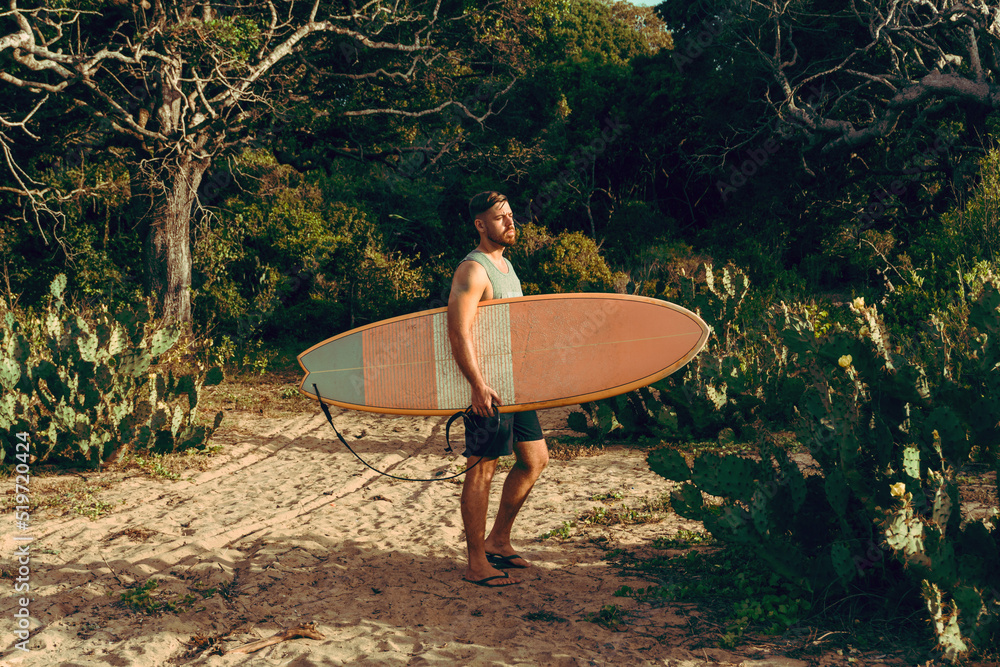 Mężczyzna surfer z deską surfingową, idący wybrzeżem na tle jungle, aktywny wypoczynek. - obrazy, fototapety, plakaty 