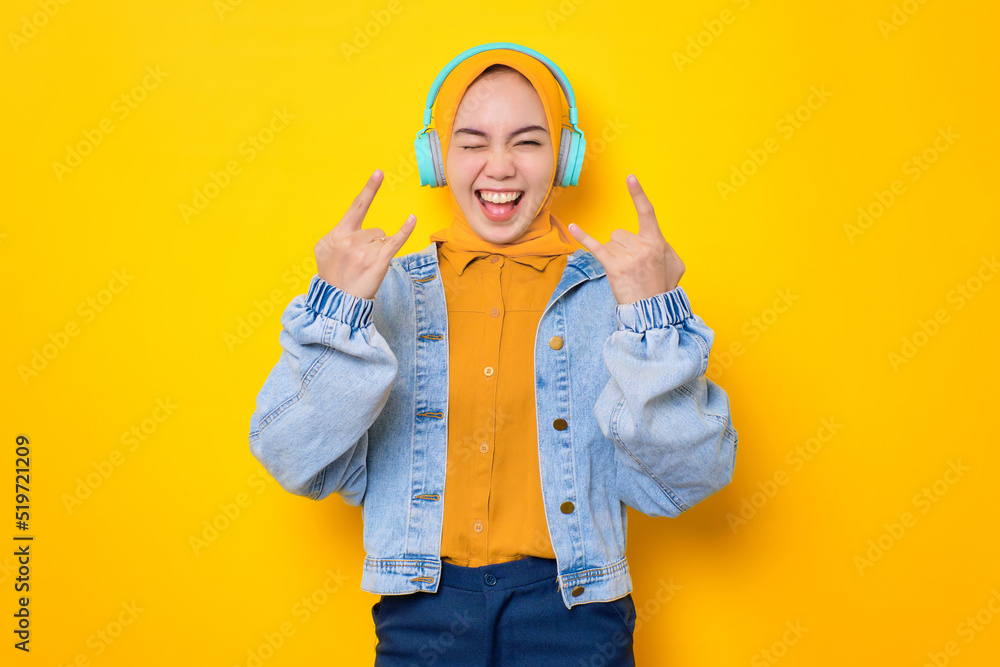 Cheerful young Asian woman in jeans jacket wearing headphones and showing rock and roll gesture isolated over yellow background Cheerful young Asian woman in jeans jacket wearing headphones and showing rock and roll gesture isolated over yellow background