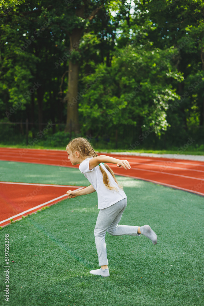 A little girl goes in for sports on the grass, the child does a warm-up before training at the stadium. Children's sports and healthy lifestyle.