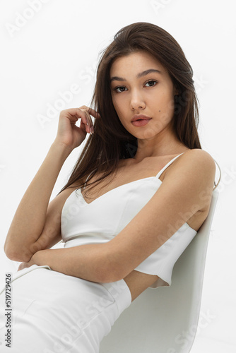 The front portrait of a young brunette woman with makeup and hairstyle, dressed in white, sits relaxed on the chair.