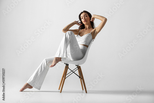 The full length image of a young brunette woman with makeup and hairstyle, dressed in white, sits relaxed on the chair.