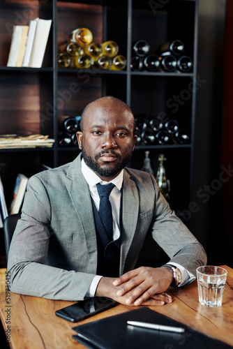 Portrait of serious African businessman in suit sitting at the table and looking at camera while waiting for somebody at restaurant.