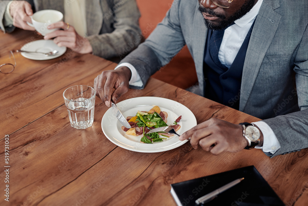 Close-up of African businessman eating salad at the table with his colleague during lunch at the restaurant Close-up of African businessman eating salad at the table with his colleague during lunch at the restaurant
