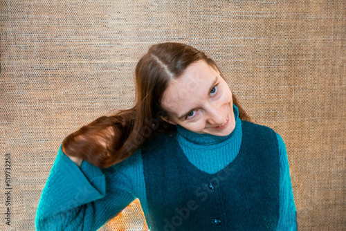 a red-haired girl in a green warm suit against a straw wall smiles and straightens her hair