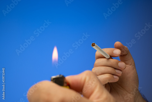 An unlit cigarette and lighter held in hand by Caucasian male hand. Close up studio shot, isolated on blue background