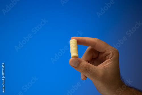 One golden corn puff held in hand by Caucasian male hand. Close up studio shot, isolated on blue background