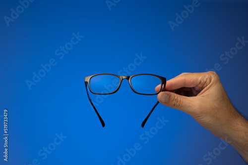 Black plastic frame eyeglasses held in hand by Caucasian male hand. Close up studio shot, isolated on blue background