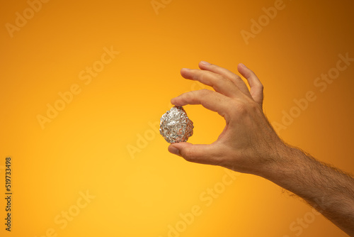 Kitchen tin foil crumpled into a ball held in hand by Caucasian male hand. Close up studio shot, isolated on orange background