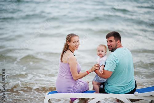Summer vacations. Parents and people outdoor activity with children. Happy family holidays. Father, pregnant mother, baby daughter sitting on sunbed at sea sand beach.