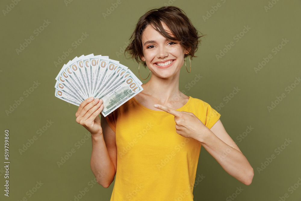 Young smiling fun satisfied happy woman she 20s wear yellow t-shirt holding point index finger on fan of cash money in dollar banknotes isolated on plain olive green khaki background studio portrait Young smiling fun satisfied happy woman she 20s wear yellow t-shirt holding point index finger on fan of cash money in dollar banknotes isolated on plain olive green khaki background studio portrait