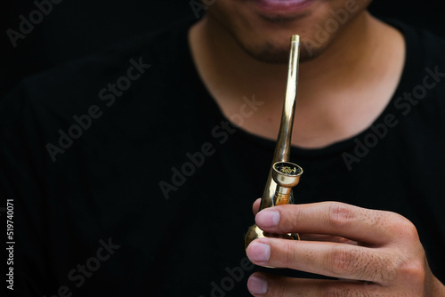 Asian man smokes marijuana from a pipe at home. Studio shoot with model simulating smoking pot with a pipe in a dark background. Cannabis legalisation.