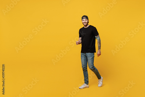 Full body young happy bearded tattooed man 20s he wears casual black t-shirt cap walking going strolling look camera isolated on plain yellow wall background studio portrait. People lifestyle concept.