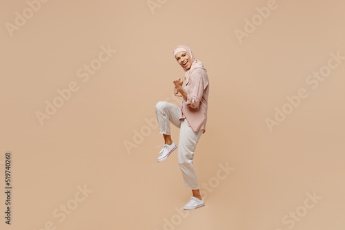 Full body young arabian asian muslim woman she wear abaya hijab pink clothes do winner gesture celebrate clenching fists say yes isolated on plain beige background. People uae islam religious concept. © ViDi Studio
