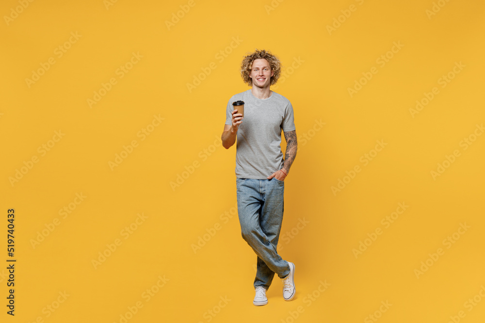 Full body young smiling man 20s he wear grey t-shirt look camera hold takeaway delivery craft paper brown cup coffee to go isolated on plain yellow backround studio portrait People lifestyle concept. Full body young smiling man 20s he wear grey t-shirt look camera hold takeaway delivery craft paper brown cup coffee to go isolated on plain yellow backround studio portrait People lifestyle concept.
