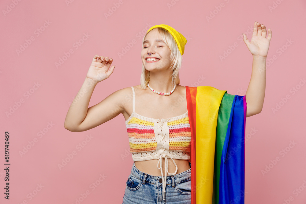 Young fun blond lesbian woman she wear colorful knitted top striped flag on shoulder dance raise up hands close eyes hat isolated on plain pastel light pink background. People lgbtq lifestyle concept. Young fun blond lesbian woman she wear colorful knitted top striped flag on shoulder dance raise up hands close eyes hat isolated on plain pastel light pink background. People lgbtq lifestyle concept.