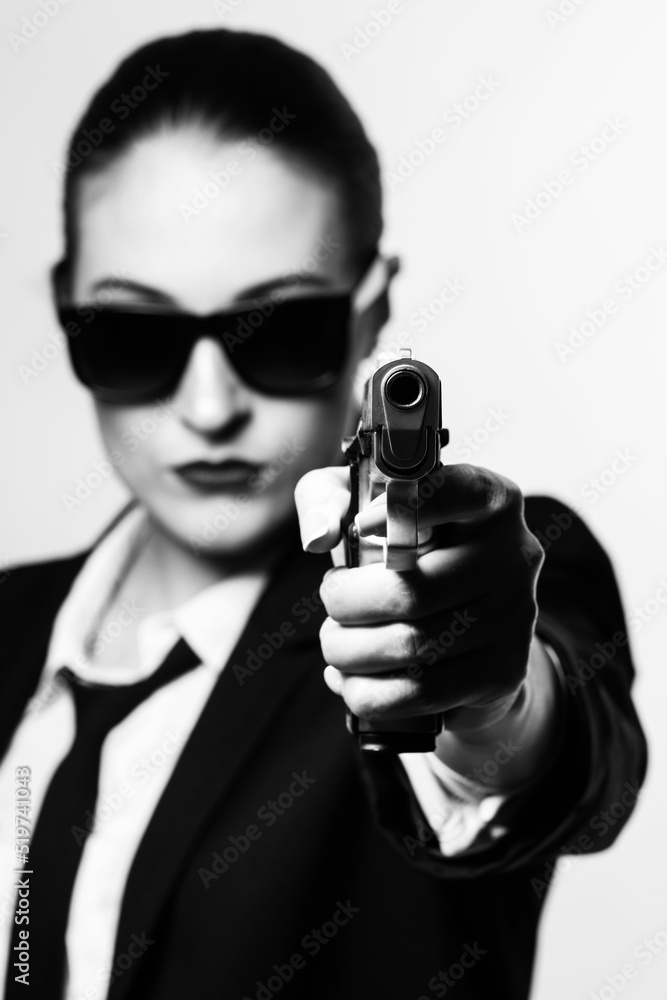 Beautiful and serious looking woman with sunglasses, classic black suit, tie and white shirt pointing pistol to camera. Gun barrel is in camera focus and model is blurred. Black and white image Beautiful and serious looking woman with sunglasses, classic black suit, tie and white shirt pointing pistol to camera. Gun barrel is in camera focus and model is blurred. Black and white image