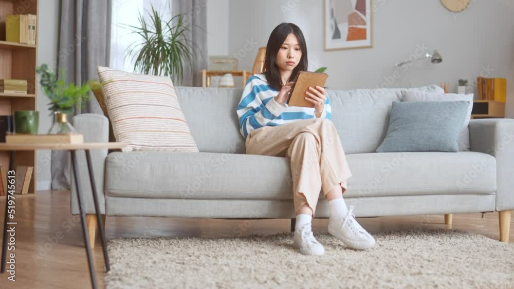 Pensive asian lady in stylish outfit surfing internet on wireless tablet while sitting on cozy sofa. Beautiful smiling woman resting at home and using modern gadget. Pensive asian lady in stylish outfit surfing internet on wireless tablet while sitting on cozy sofa. Beautiful smiling woman resting at home and using modern gadget.