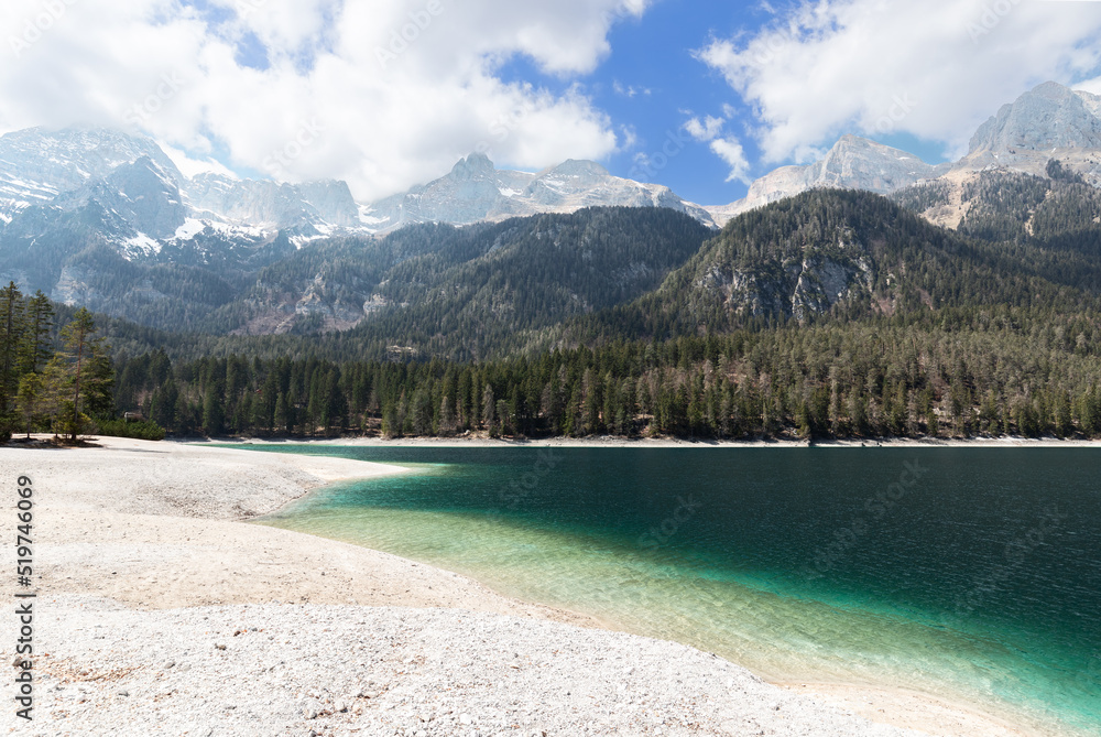 Panorama of the gentle shore of Tovel lake with emerald water and alpine mountain ranges covered with forest and snow in the background. Ville d'Anaunia, Trentino, Italy