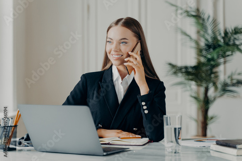 Smiling young attractive businesswoman talking on phone in front of laptop