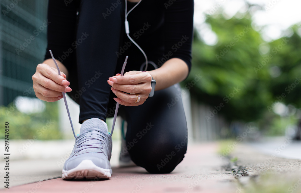 Crop unrecognizable woman runner wearing hygiene protective face mask sitting and tieing shoe rope during workout running in city park. Concept of healthcare in time of coronavirus outbreak