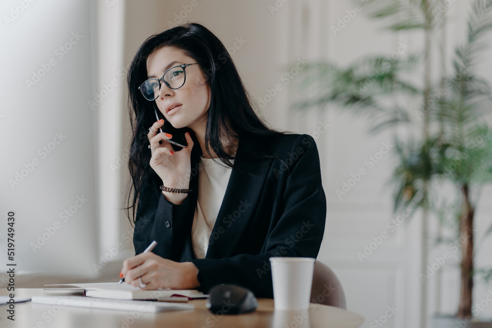 Gorgeous businesswoman makes phone call, concentrated in computer screen, writes down information into notepad, poses at cozy workplace, focused on important task, makes notes in personal organizer Gorgeous businesswoman makes phone call, concentrated in computer screen, writes down information into notepad, poses at cozy workplace, focused on important task, makes notes in personal organizer