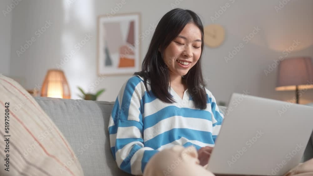 Young focused woman sitting on sofa and working on modern laptop in cozy apartment. Smiling asian female looking thoughtful while writing work email.