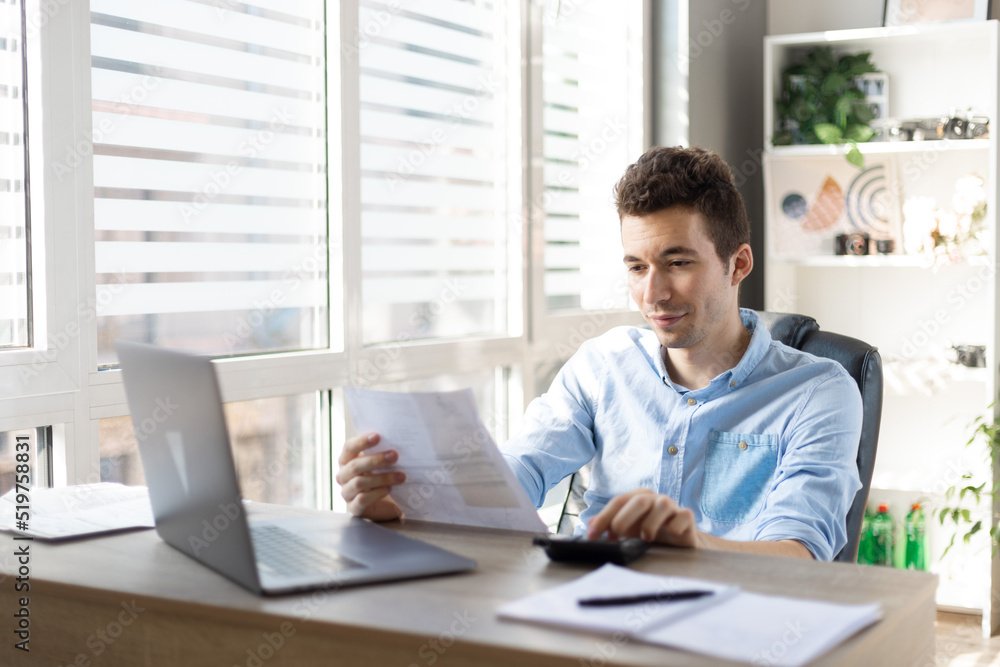Young businessman checking bills, holding receipt, using calculator, accountant calculating company expenses, financial report, sitting at wooden work desk, young male planning budget Young businessman checking bills, holding receipt, using calculator, accountant calculating company expenses, financial report, sitting at wooden work desk, young male planning budget