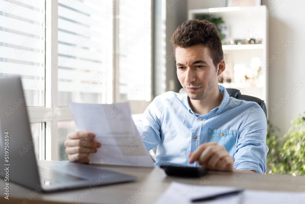 Young businessman checking bills, holding receipt, using calculator, accountant calculating company expenses, financial report, sitting at wooden work desk, young male planning budget