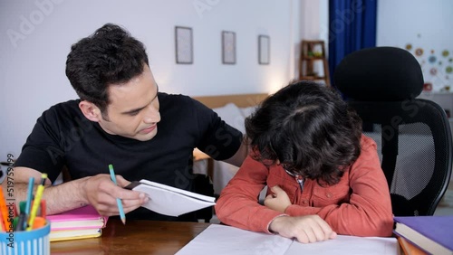 An Indian father scolding his young son as he puts his head down and cries - an angry father and a depressed little kid. An Indian father is angry with his young son over his study performance - sc... photo