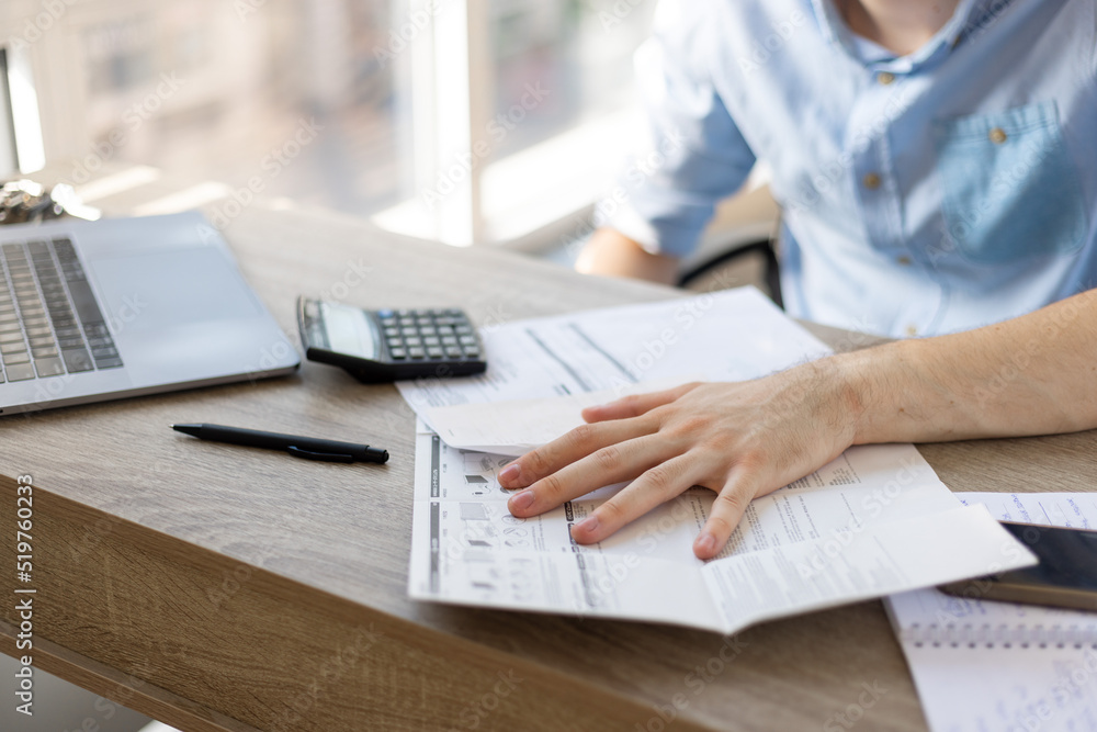Close up young man using calculator summarizing domestic utility bills or taxes, managing household monthly budget, planning investment, reviewing financial payments alone at home, using computer app. Close up young man using calculator summarizing domestic utility bills or taxes, managing household monthly budget, planning investment, reviewing financial payments alone at home, using computer app.