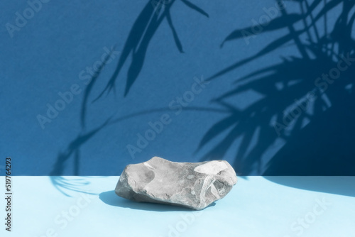 The podium is made of gray stone on a blue background with a shadow from the leaves of a palm tree. Scene for product promotion, beauty, natural eco cosmetic. Showcase, display case.