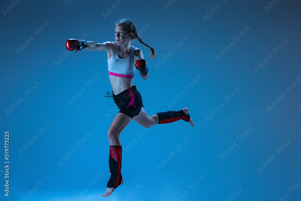 Dynamic portrait of young girl, junior MMA fighter in action, motion isolated on blue background in neon light. Concept of kids sport