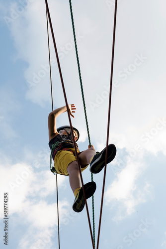 Child in safety harness and helmet in rope park. Boy climbs attached with carbine to cable on rope way.