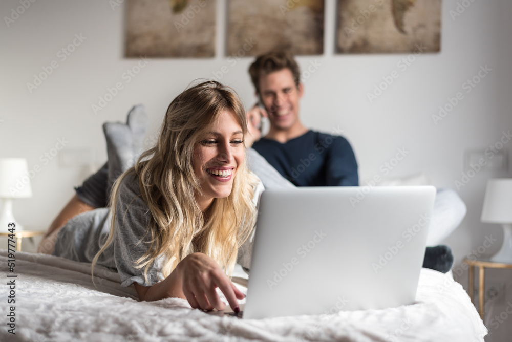 young couple on bed entertained on their computer and phone young couple on bed entertained on their computer and phone