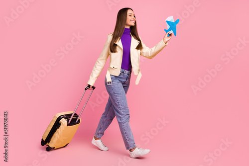 Full length profile photo of cute young lady with valise hold plane look promotion wear jacket jeans boots isolated on pink color background