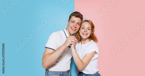 Portrait of young cheerful couple, singing in microphone isolated over pink blue studio background. Karaoke birthday celebration © Lustre Art Group 