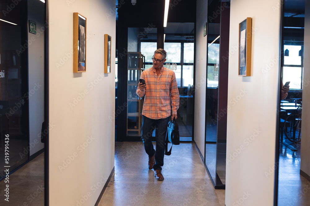 Caucasian mature businessman using smart phone while walking in corridor of creative office