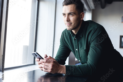 Thoughtful caucasian young businessman with smart phone looking through window in office